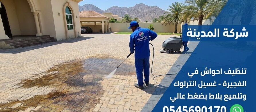 فريق عمل شركة تنظيف احواش في الفجيرة يستخدم ماكينة ضغط عالي وماكينة جلي لتنظيف وإزالة البقع من أرضية انترلوك واسعة في فيلا، مع ظهور جبال الفجيرة في الخلفية.