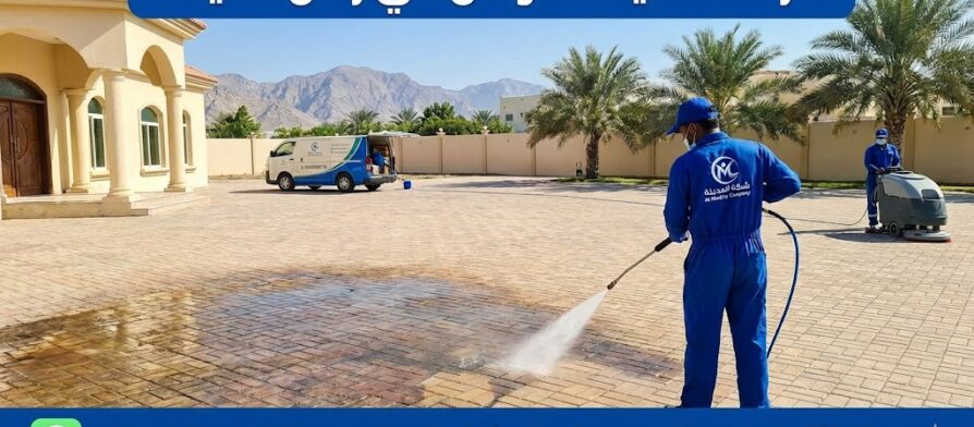 عمال شركة تنظيف احواش في راس الخيمة يستخدمون ماكينات الغسيل بالضغط العالي لتنظيف انترلوك فيلا واسعة وإزالة البقع، مع ظهور جبال راس الخيمة في الخلفية
