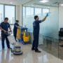فريق عمل شركة تنظيف مكاتب في عجمان يقوم بتلميع الأرضيات وتنظيف الفواصل الزجاجية وكنس السجاد داخل مقر إداري حديث.