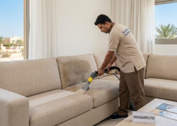 فني محترف من شركة تنظيف كنب في الظفرة يرتدي زي شركة المدينة ويستخدم ماكينة التنظيف بالحقن والاستخلاص لإزالة البقع من كنب بيج كبير في صالة منزل فاخر تطل نوافذه على أشجار النخيل.