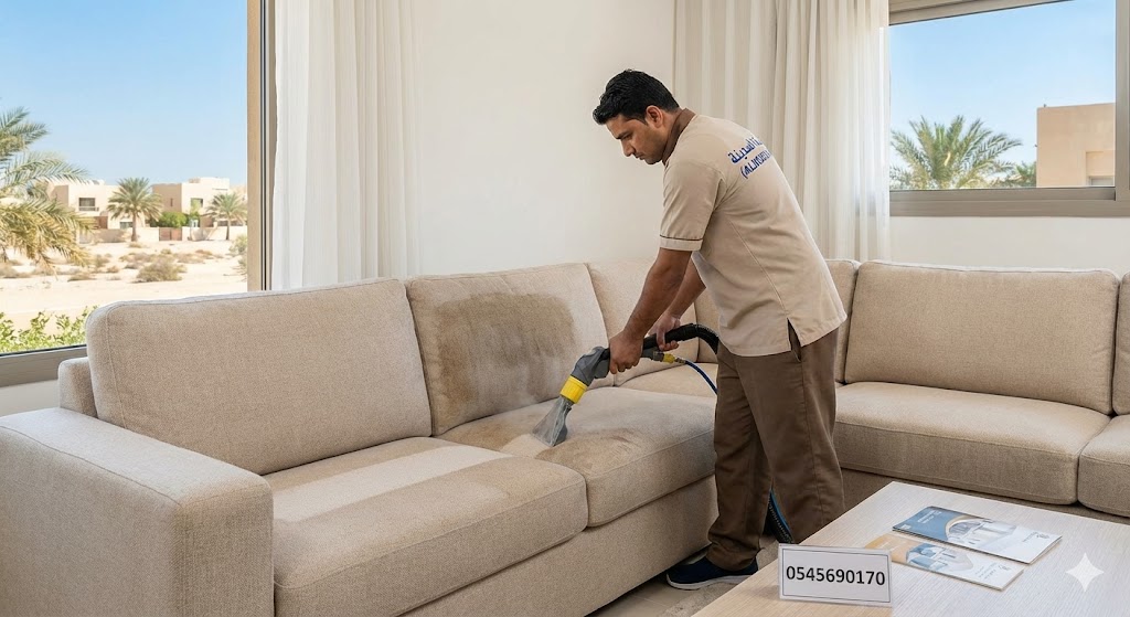 فني محترف من شركة تنظيف كنب في الظفرة يرتدي زي شركة المدينة ويستخدم ماكينة التنظيف بالحقن والاستخلاص لإزالة البقع من كنب بيج كبير في صالة منزل فاخر تطل نوافذه على أشجار النخيل.