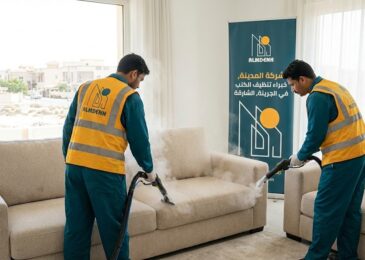 النص البديل عمال شركة المدينة، بصفتها شركة تنظيف كنب في الجرينة بالشارقة، يقومون بتنظيف كنب فاتح اللون باستخدام تقنية البخار داخل فيلا، مع ظهور لافتة إعلانية تحمل الهوية البصرية للشركة.