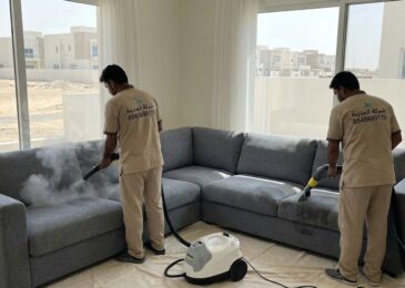 فنيان من شركة المدينة يستخدمان أجهزة التنظيف بالبخار والشفط لغسيل كنب زاوية كبير داخل فيلا حديثة مطلة على مناطق مفتوحة، مما يوضح احترافية خدمات أفضل شركة تنظيف كنب في الرقايب.