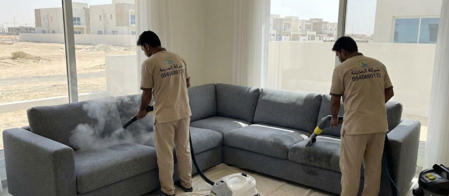 فنيان من شركة المدينة يستخدمان أجهزة التنظيف بالبخار والشفط لغسيل كنب زاوية كبير داخل فيلا حديثة مطلة على مناطق مفتوحة، مما يوضح احترافية خدمات أفضل شركة تنظيف كنب في الرقايب.