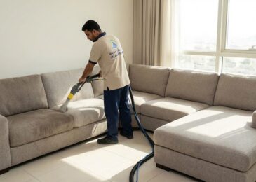 فني متخصص من شركة تنظيف كنب في الروضة يستخدم جهاز الحقن والاستخلاص لتنظيف كنب قماش رمادي كبير في صالة منزل حديث بالشارقة مع ظهور شعار شركة المدينة ورقم التواصل.