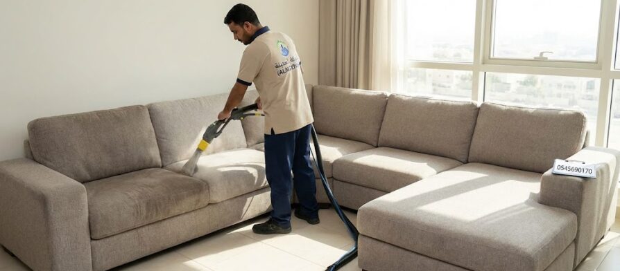 فني متخصص من شركة تنظيف كنب في الروضة يستخدم جهاز الحقن والاستخلاص لتنظيف كنب قماش رمادي كبير في صالة منزل حديث بالشارقة مع ظهور شعار شركة المدينة ورقم التواصل.