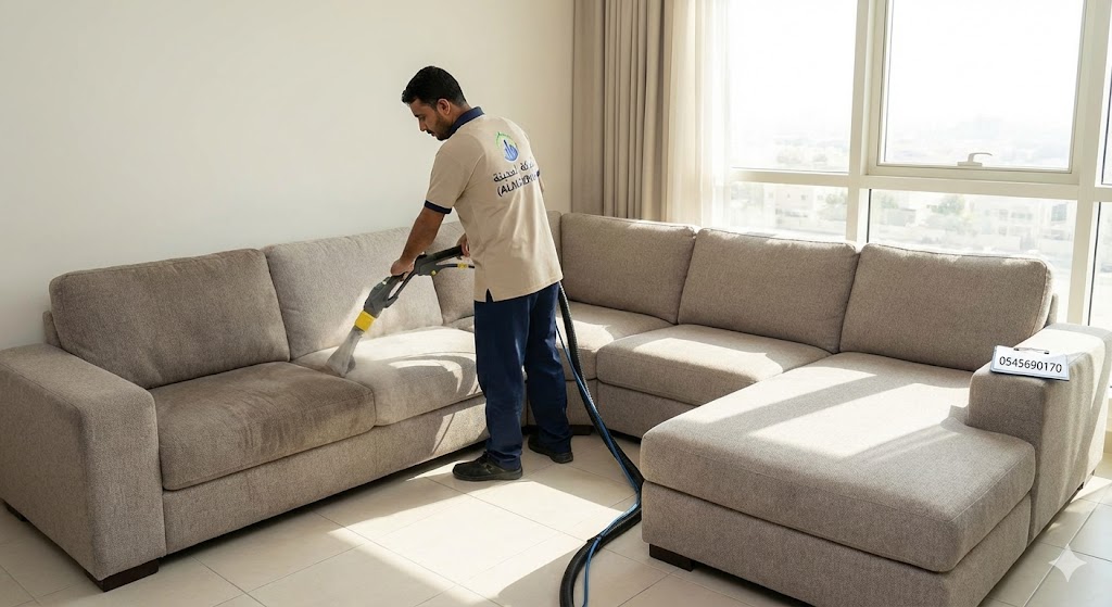 فني متخصص من شركة تنظيف كنب في الروضة يستخدم جهاز الحقن والاستخلاص لتنظيف كنب قماش رمادي كبير في صالة منزل حديث بالشارقة مع ظهور شعار شركة المدينة ورقم التواصل.
