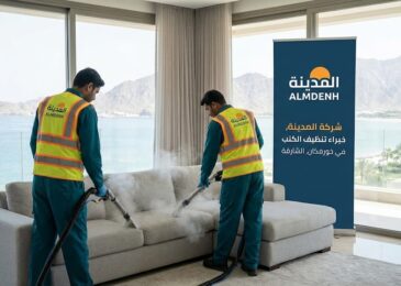 عمال شركة المدينة، بصفتها شركة تنظيف كنب في خورفكان، يقومون بتنظيف كنب فاتح اللون بالبخار في غرفة ذات إطلالة بانورامية على البحر والجبال، بجوار لافتة تحمل شعار الشركة.