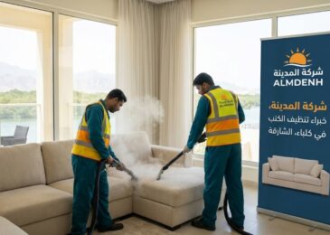 عمال شركة المدينة، بصفتها شركة تنظيف كنب في كلباء، يقومون بغسيل كنب فاتح اللون بالبخار في غرفة ذات إطلالة طبيعية خلابة، بجوار لافتة تحمل شعار الشركة.