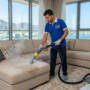 فني متخصص من شركة تنظيف كنب في الفجيرة يستخدم آلة استخراج البخار لتنظيف أريكة قماشية حديثة بيج بعمق، مع إطلالة بانورامية على جبال الحجر والبحر في الفجيرة.