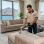 فني محترف يستخدم آلة تنظيف عميق على أريكة رمادية حديثة في غرفة معيشة فاخرة، مع إطلالة بانورامية على الجبال والبحر في الفجيرة، كجزء من خدمات شركات تنظيف الكنب في الفجيرة.